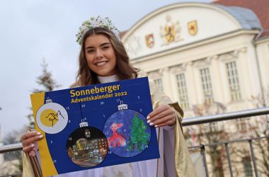 Eine Frau ist als Christkind gekleidet und hält den Sonneberger Adventskalender 2022 in den Händen. Im Hintergrund ist das Rathaus der Stadt Sonneberg.
