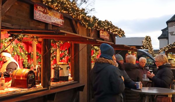 Ein Verkaufsstand mit der Aufschrift: Müllers Glühweinschanke. Vor dem Stand stehen Menschen an Stehtischen.