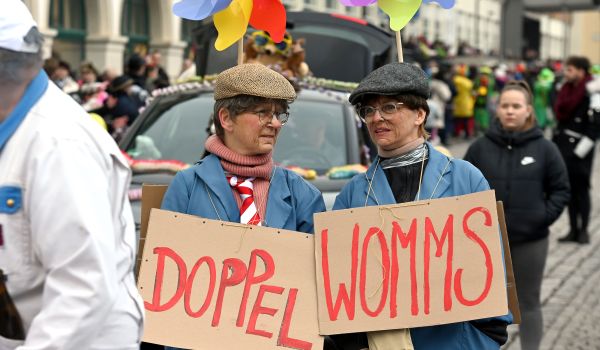 Zwei Frauen mit blauem Mantel und Schiebermütze. Sie tragen Schilder mit der Aufschrift: Doppel Womms um den Halz.