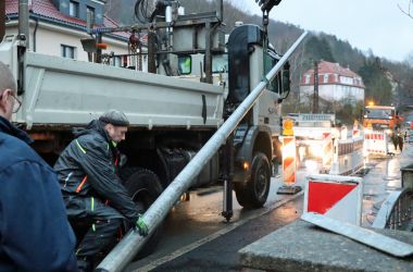 Zwei Bauarbeiter setzen einen Lichtmast auf einem Gehweg.