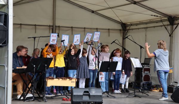 Kinder stehen auf einer Bühne unter einem weißen Zelt. Eine Frau steht vor ihnen und dirigiert.