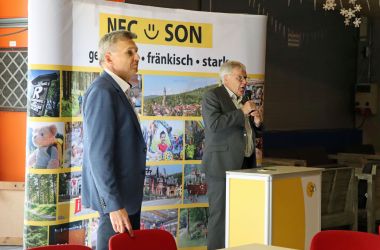 Ein Mann mit Mikrofon steht an einem Rednerpult. Es ist Peter Soyer. Links daneben steht ein weiterer Mann. Es ist der Bürgermeister Dr. Heiko Voigt. Hinter den beiden steht ein Banner mit der Aufschrift: NEC - SON; gemeinsam, fränkisch, stark.