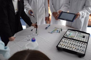 Auf einem Tische liegt ein Experimentierkoffer. Dahinter stehen Menschen in weißen Kitteln.