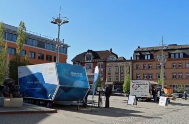 Ein blau-weißer Anhänger steht auf einem Platz, umrahmt von Häusern.