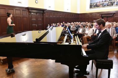 Ein Mann sitzt an einem Flügel und spielt. Hinter ihm sitzt ein Publikum.