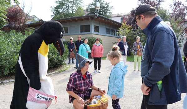 Im Stadtpark: Ein Mann mit spitzem Hut hockert sich vor ein kleines Mädchen. Vor ihm steht ein Korb mit Geschneken. Daneben steht eine Person im Pinguin-Kostüm. Es trägt die Aufschrift: SonneBad.