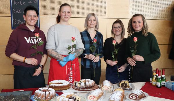 Fünf Frauen stehen hinter einem Tisch und halten eine Rose in der Hand. Auf dem Tisch stehen verschiedene Kuchen.