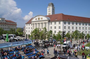 Vor dem Rathaus der Stadt Sonneberg stehen viele Autos. Im unteren Teil des Bildes steht eine Tribühne mit vielen Menschen darauf.