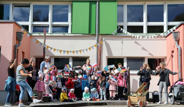 Kinder und Erwachsene stehen vor einem bunten Gebäude und halten Flaggen auf verschiedenen Ländern in den Händen.