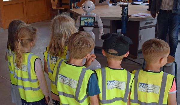 Kinder mit Warnweste stehen vor einem Roboter und schauen ihn an.