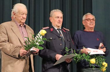 Drei ältere Männer stehen mit einer Urkunde und Blumen in der Hand vor einem dunklen Vorhang.