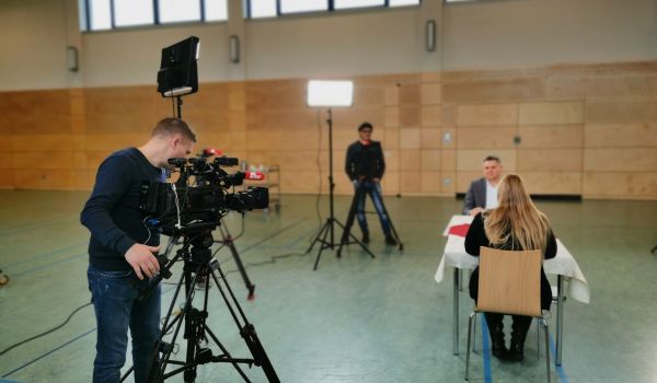 An einem Tisch sitzen zwei Personen. Daneben steht ein Kamerateam und filmt.