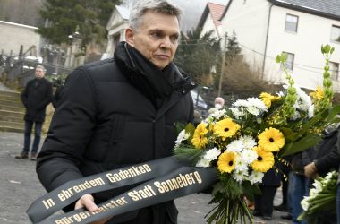 Ein schwarz gekleideter Mann hält einen Blumenestrauß mit Trauerband.