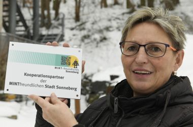 Eine Frau hält ein Schild in der Hand. Das Schild trägt die Aufschrift: Kooperationspartner der Mintfreundlichen Stadt Sonneberg.