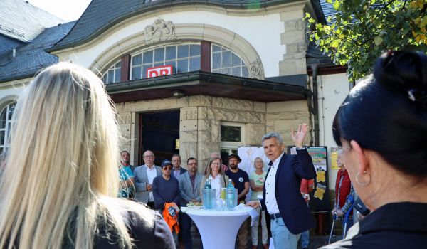 Einige Menschen stehen vor dem Bahnhofsgebäude um einen Stehtisch.