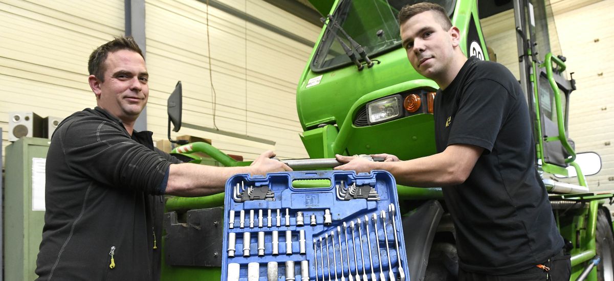 Zwei Männer mit Werkzeukasten stehen vor einer Landwirtschaftmaschine.