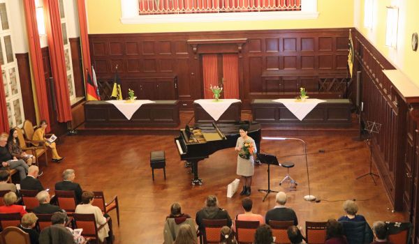 Eine Frau steht neben einem Flügel und hält einen Blumenstrauß in den Händen. Vor ihr sitzen viele Menschen.