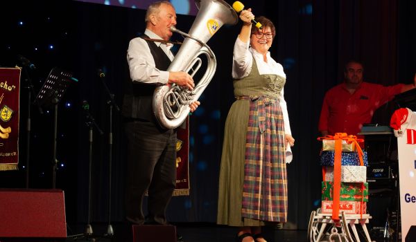 Ein Mann und eine Frau stehen auf einer Bühne. Der Mann spielt Tuba. Die Frau hält ein Mikrofon in der Hand.