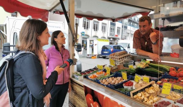 Zwei Frauen stehen vor einem Verkaufsauto und interviewen den Verkäufer mit einem Mikrofon.