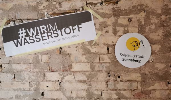 An einer Rohbauwand hängt eine Aufkleber mit der Aufschrift: Wir im Wasserstoff. Daneben hängt ein weiterer mit Spielzeugstadt Sonneberg.