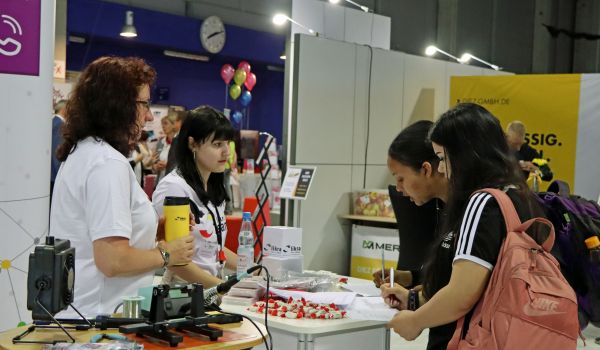 Zwei Frauen stehen hinter einem Werbestand. Auf dem Stand liegen Werbematerialien. Zwei Jugendliche stehen davor und füllen einen Zettel aus.