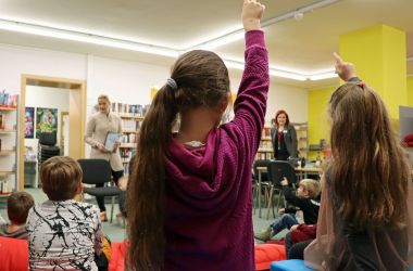 Kinder sitzen in einem Kreis. Drei Kinder melden sich. Vor ihnen steht eine Frau mit einem Buch in der Hand.