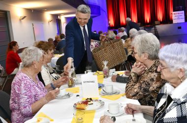 Senioren sitzen an gedeckten Kaffeetafeln in einem großen Saal. Ein Mann verteilt Geschenke.