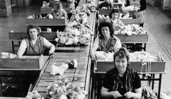 Schwarz-weiß: Frauen sitzen in einer Fabrikhalle am Fließband. Auf dem Fließband liegen Plüschtiere.