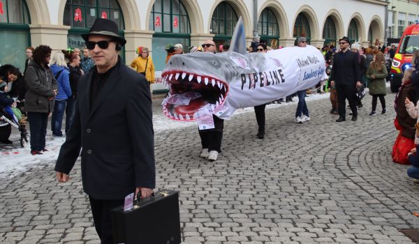 Drei Menschen tragen ein gemeinsames Haikostüm mit der Aufschrift Pipeline. Im Maul des Haies sind Geldscheine.