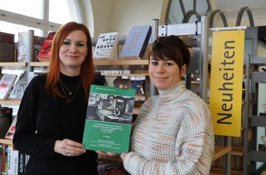 Zwei Frauen halten in einer Bibliothek eine A4-große Publikation in die Kamera.