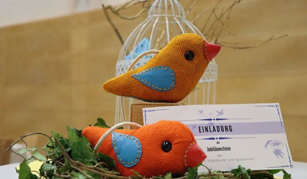 Auf einem Tisch steht eine Nest mit zwei bunten Deko-Vögeln aus gewebtem Stoff. Daneben ist eine Karte mit der Aufschrift: Einladung zur Jubiläumsfeier.