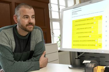Ein Mann sitzt vor einem Monitor. Der Monitor zeigt das Portal der Online-Terminvergabe.