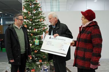 Ein Mann und eine Frau überreichen einen Scheck. Im Hintergrund steht ein geschmückter Weihnachtsbaum.