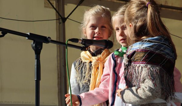 Drei Kinder in Tracht stehen vor einem Mikrofon auf einer Bühne. Im Vordergrund liegt Gemüse auf einem Tisch.