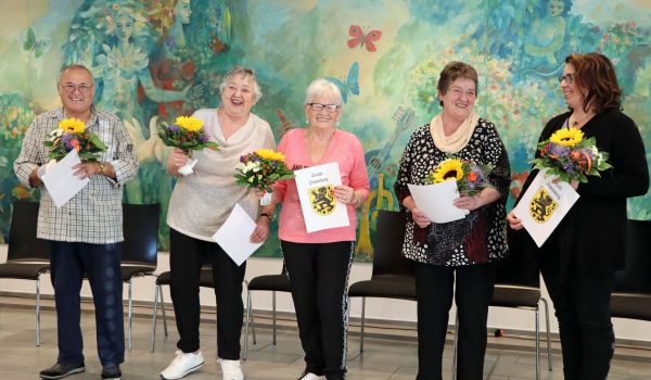 Fünf Personen stehen in einer Reihe nebeneinander. Sie halten eine Urkunde und Blumen in den Händen.