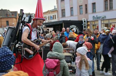 Ein kostümierter Mann, mit rotem Rock und spitzem Hut, spielt Gitarre. Um ihn herum befinden sich viele Menschen.