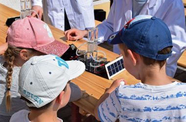Drei Kinder stehen an einem Tisch. Auf dem Tisch steht eine Brennstoffzelle zum Experimentieren mit Wasserstoff.