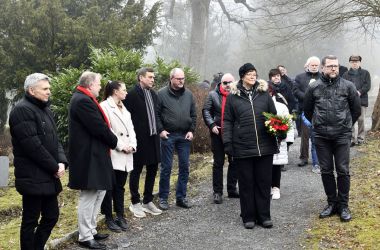 Mehrere schwarzgekleidete Personen bringen Blumen auf einen Friedhof.