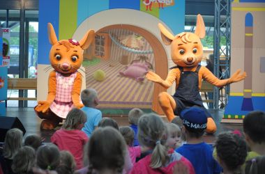 Zwei Mausfiguren stehen auf einer Bühne und davor stehen Kinder.