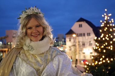 Eine junge Frau mit blonden Locken trägt ein Christkind-Kostüm und befindet sich auf dem Rathausbalkon mit dem Weihnachtsbaum im Hintergrund.