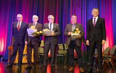 Fünf Männer stehen auf einer Bühne. Die drei mittleren halten Präsente und Urkunden in der Hand. Links davon steht Professor Dippold und rechts der Bürgermeister Dr. Voigt.