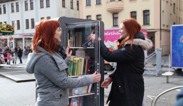 Zwei Menschen stellen ein Buch in den Bücherschrank. Dieser befindet sich auf einem Marktplatz.