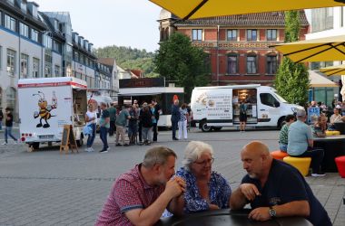 Zwei Männer und eine Frau sitzen an einem Tisch auf einem Marktplatz und unterhalten sich.