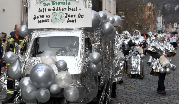 Ein Auto ist in silberne Folie gewickelt und mit Luftballons geschmückt. Dahinter laufen Menschen, die auch in Folie gewickelt sind.