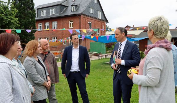 Eine Gruppe Männer und Frauen steht im Halbkreis auf einer Wiese vor einem Backsteinhaus.