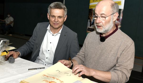 Zwei Männer sitzen an einem Tisch. Der Rechte blättert in einem Buch.