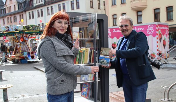 Zwei Menschen stellen ein Buch in den Bücherschrank. Dieser befindet sich auf einem Marktplatz.
