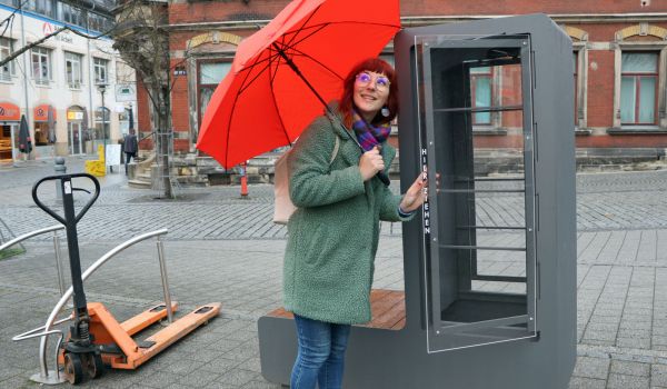 Ein Frau mit rotem Regenschirm steht vor einem Bücherschrank.