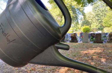 Eine Gießkanne mit der Aufschrift Friedhof hängt an einem Metallgerüst vor einer Grabstelle.