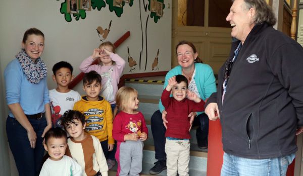 Kinder und Erwachsene stehen in einem Treppenaufgang. Im Hintergrund ist eine mit Bäumen bemalte Wand.